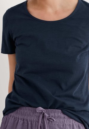 Personne portant un t-shirt à manches courtes et encolure ronde de couleur bleu marine foncé, rentré dans un pantalon à taille élastique violet clair avec un nœud à l'avant.