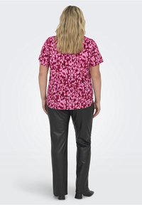 Kobieta z długimi blond włosami, ubrana w różową bluzkę z krótkim rękawem w wzory oraz czarne spodnie o prostym kroju, stoi przodem do widza.