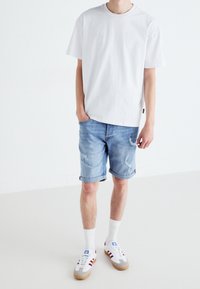 Bílá bavlněná tričko, světle modré džíny s vybledlými detaily, bílé ponožky a šedé tenisky s červenými pruhy a hnědými podrážkami.