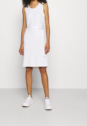 Mujer con vestido blanco sin mangas hasta la rodilla con un cinturón delgado y zapatillas blancas con plataforma, de pie contra un fondo liso y claro.
