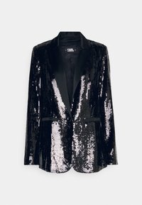 Blazer negro con lentejuelas y solapas de satén, cierre de un botón y dos bolsillos frontales. Las lentejuelas iridiscentes reflejan la luz desde diferentes ángulos.
