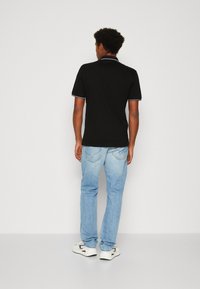 Camisa polo negra con cuello y acentos en las mangas blancos, combinada con jeans azul claro. El modelo está usando zapatillas blancas y está de espaldas.