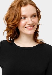 Zwarte geribbelde lange mouwen top met een ronde halslijn, met een gestructureerde stof. Het model heeft golvend rood haar en een neutrale achtergrond.