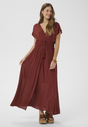 Femme portant une robe maxi fluide de couleur rouille orangée avec des manches courtes et une taille cintrée, associée à des sandales à plateforme et des bijoux dorés.