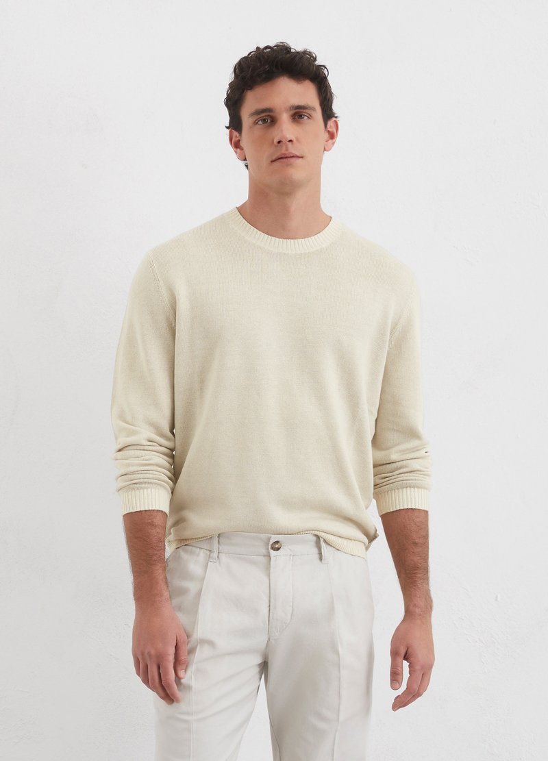 Cremfarbener Pullover mit Rundhalsausschnitt, langen Ärmeln und gerippten Bündchen, kombiniert mit hellen Hosen, vor einem schlichten weißen Hintergrund.