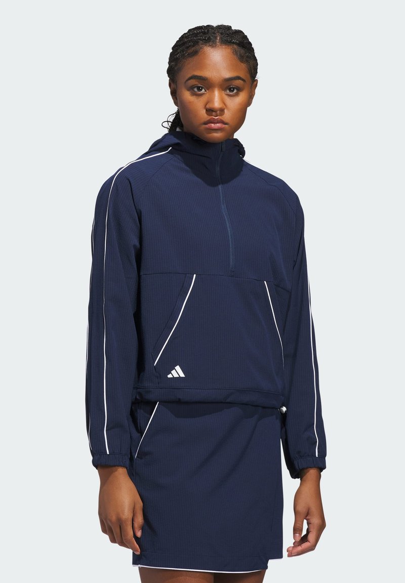 Námornícka modrá športová bunda s polovičným zipsom, rebrovanou textúrou, bielymi akcentmi a elastickými manžetami. Má štvrťzipsový golier a bočné vrecká.