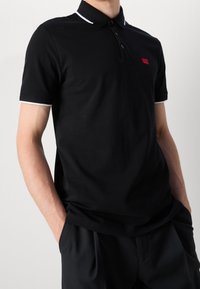 Svart pikétröja i bomullsblandning, med ribbat krage och vita detaljer, tre-knappars knäppning och en liten r öd logotyp på bröstet.