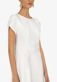 Robe blanche avec des manches courtes légèrement bouffantes, dotée d'un détail de taille en dentelle florale et d'une texture satin lisse. Design élégant, style minimaliste.