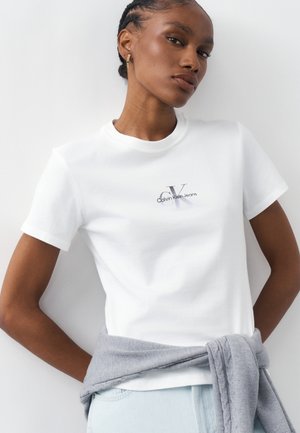 Γυναίκα που φοράει λευκό t-shirt Calvin Klein Jeans, ανοιχτόχρωμο τζιν και γκρι πουλόβερ δεμένο γύρω από τη μέση, μπροστά σε απλό λευκό φόντο.