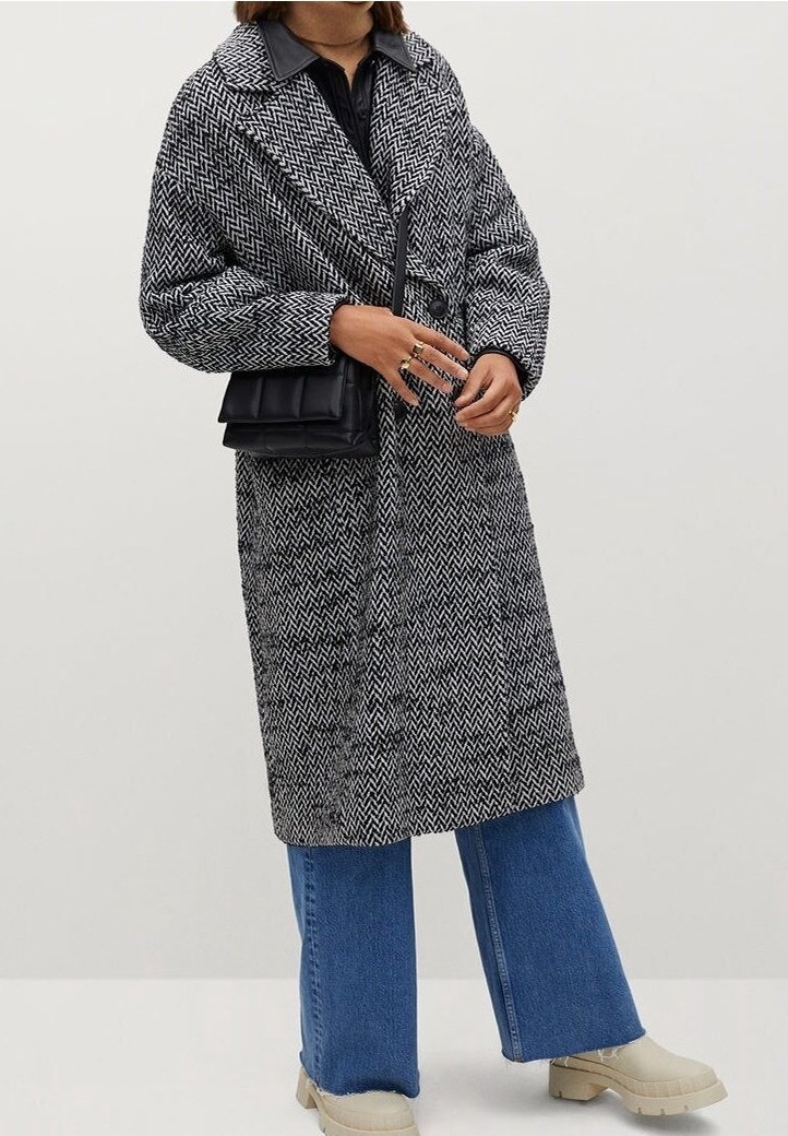 Person, der einen schwarz-weißen Fischgrätenmantel, blaue weite Jeans, beigen klobigen Schuhen trägt und eine kleine schwarze Umhängetasche bei sich hat.