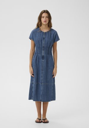 Moteris stovinti, dėvinti vidutinio ilgio, trumpomis rankovėmis mėlyną džinsinę suknelę su priekiniais užtrauktuku, klostuotu liemeniu ir juodais sandalais ant balto fono.