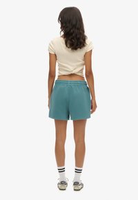Shorts de algodón azul verdoso con cinturilla elástica, que presentan un corte holgado y una textura suave. Combinados con una parte superior ajustada en color crema.