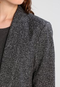 Blazer grigio con texture, realizzato in un morbido tessuto intrecciato con un motivo sottile, ampie rever e una vestibilità sartoriale sulle spalle.