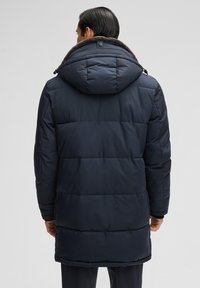 Navyblauwe puffermantel met een capuchon, voorzien van horizontale gestikte panelen, een met bont gevoerde kraag en drukknoopdetails bij de hals.