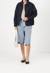 Marineblauwe jas, beige T-shirt, lichtblauwe denim shorts, zwarte platte schoenen en een kleine beige tas. Casual outfit met eenvoudige, strakke lijnen.