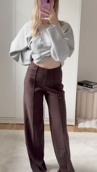 Harmaa lyhythihainen collegepaita, jossa on vetoketju edessä ja leveät hihat, yhdistettynä korkeavyötäröisiin tummanruskeisiin housuihin, joissa on keskisaumat.