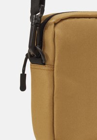 Beige stoffen schoudertas met een gestructureerd oppervlak, voorzien van een ritssluiting en zwarte hardware-accenten, waaronder een stevige haak en een trekband.