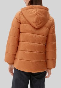 Kobieta w kapturze, ubrana w pikowaną, pomarańczowo-brązową kurtkę puchową i ciemne spodnie, widoczna z tyłu na jednolitym szarym tle.