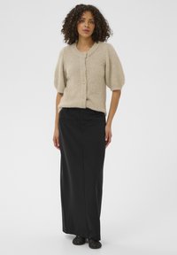 Cardigan en tricot beige à manches courtes, col rond et fermeture à boutons, associé à une longue jupe noire et des chaussures sombres.