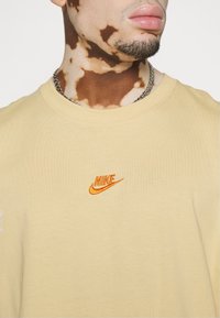 Beżowa bawełniana koszulka z okrągłym dekoltem i krótkimi rękawami. Posiada haftowane pomarańczowe logo Nike na piersi.