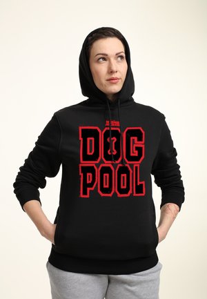 Kvinde iført en sort hættetrøje med rød tekst "Dog Pool" og grå joggingbukser, står med hænderne i lommerne og kigger til siden.