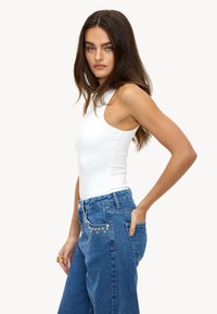 Body blanc moulant avec un col montant et des bretelles type débardeur, associé à un jean denim bleu clair orné de clous et à la coupe décontractée.