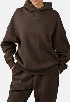 Donna che indossa una felpa con cappuccio oversize marrone scuro e pantaloni della tuta coordinati con le mani in tasca su uno sfondo semplice.