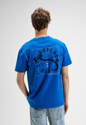 Man met krullend haar die een blauw T-shirt draagt met de tekst "BUTCHER OF BLUE" en een zwarte vleeshaakgrafiek op de rug, gecombineerd met lichte jeans.