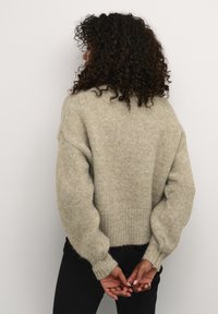 Pull en maille oversized gris clair avec une finition texturée, des poignets côtelés et un ourlet arrondi. Le mannequin le porte avec un pantalon noir, les mains jointes derrière.