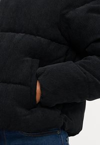 Svart corduroy puffjakke med en strukturert overflate, med brede horisontale paneler og sidelommer for oppbevaring.