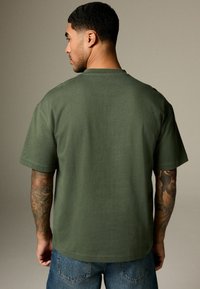 Olive green T-shirt with short sleeves, round neckline, and a smooth texture; worn with blue denim jeans, displaying tattooed forearm.