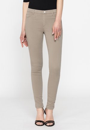 Pantaloni beige slim-fit indossati con top nero e tacchi neri con cinturini e punta aperta, mostrati dalla vita ai piedi su sfondo bianco.