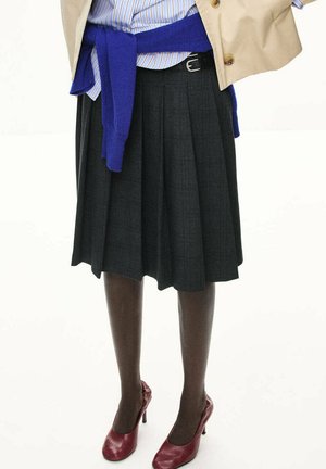 Persona que lleva una falda plisada gris oscuro, medias negras, tacones altos rojos, un suéter azul atado a la cintura, camisa de rayas y abrigo beige.