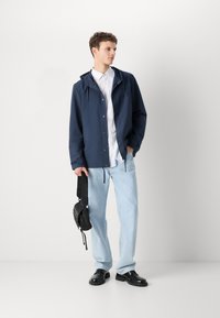 Navy waterproof jacket with a hood, white button-up shirt, light blue jeans, and black shoes. Model holds a small black crossbody bag.