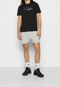 Černé tričko s krátkým rukávem a vícebarevným logem, šedé kraťasy a černé tenisky. Doplňky zahrnují bílé ponožky s detailním logem.