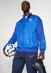 Modrá bunda na zips s vyrazenými písmenami, bielym logom Nike, doplnená tmavomodrými nohavicami. Hráč drží bielu futbalovú loptu a čierne kopačky.