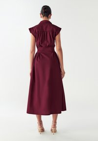 Abito borgogna lungo fino alla caviglia con maniche a cappuccio, vita aderente e schiena plissettata. Tessuto morbido, design minimalista e chiusura con zip invisibile.