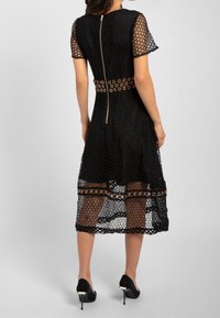 Robe en dentelle noire avec manches courtes, découpes circulaires, taille ajustée avec ceinture à œillets dorés et fermeture éclair dissimulée au dos.