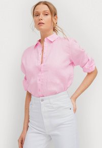 Camicia di lino rosa chiaro con bottoni, maniche arrotolate e colletto, abbinata a jeans bianchi a vita alta.