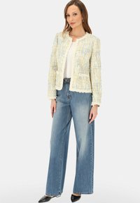 Kobieta w kremowej, teksturowanej kurtce z odkrytym przodem, białej bluzce, szerokich, wyblakłych niebieskich dżinsach i czarnych, szpiczastych butach, stojąca na jednolitym tle.