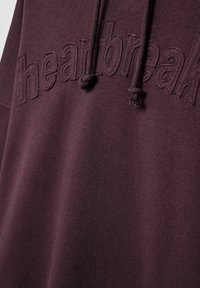 Ombra jopica s kapuco bordo barve z vezenim napisom "heartbreak" v ujemajoči se niti. Ima kapuco z vrvico in mehak, teksturiran material.