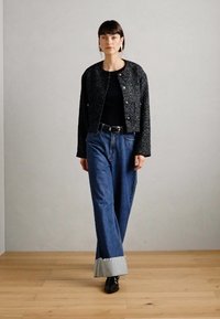 Veste noire texturée courte avec des boutons métalliques, associée à un jean bleu taille haute avec des ourlets pliés bleu clair et des bottines noires.