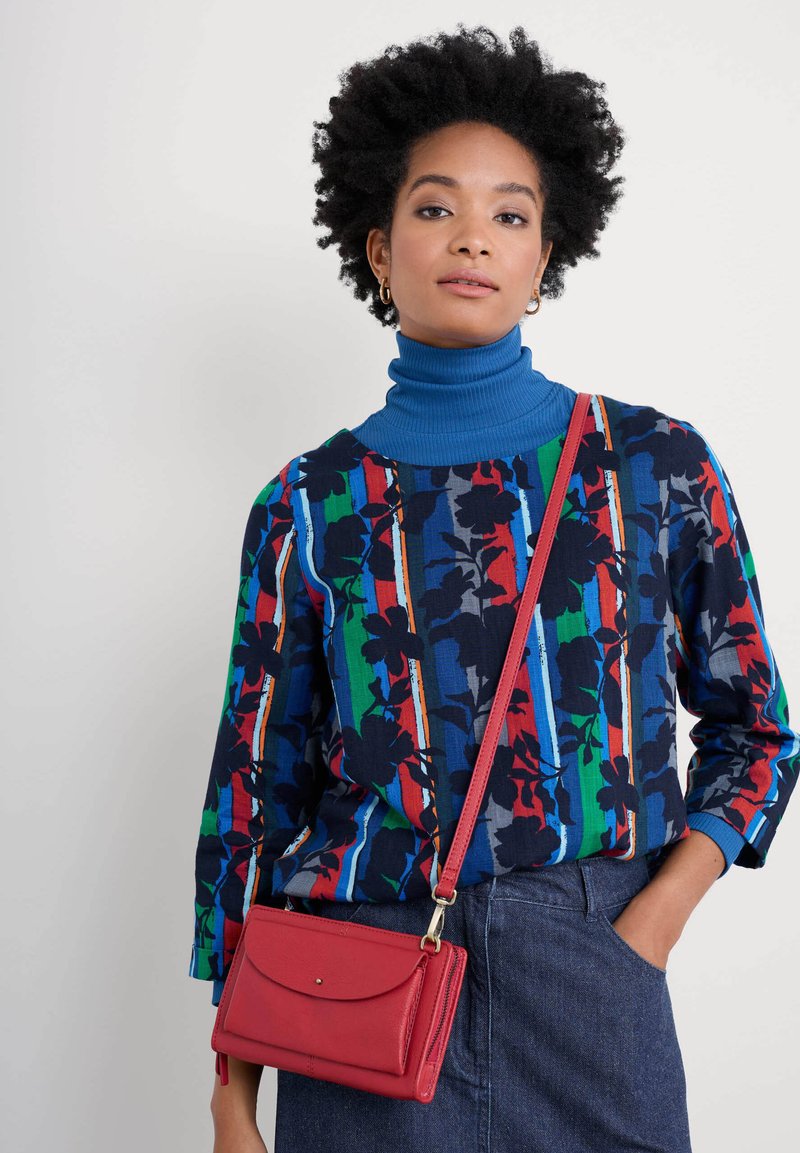 Bunt gemusteter Pullover mit blauem Rollkragen, kombiniert mit einer roten Leder-Crossbody-Tasche, einem Jeansrock und strukturiertem schwarzen Haar.