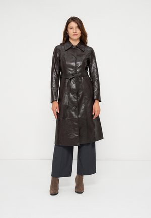 Femme debout portant un long manteau en cuir noir brillant avec boutons et ceinture, un pantalon large et sombre, et des bottines en daim marron sur fond blanc.
