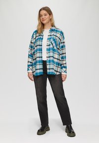 Camicia in flanella a quadri blu e bianchi sopra una maglietta bianca, abbinata a jeans neri e stivali neri massicci. Fit rilassato con dettaglio della tasca.
