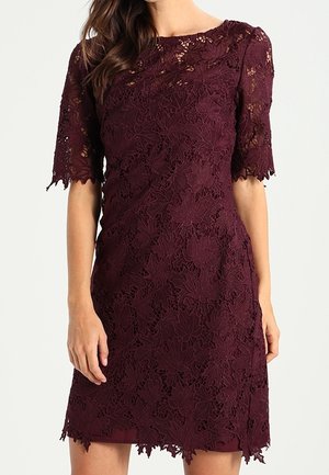 Femme portant une robe en dentelle violet foncé, courte aux manches et longueur genou, avec des motifs floraux et des bords festonnés aux manches et à l’ourlet.