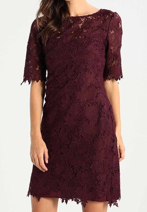Femme portant une robe en dentelle violet foncé, courte aux manches et longueur genou, avec des motifs floraux et des bords festonnés aux manches et à l’ourlet.