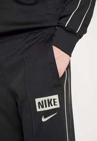 Černé tepláky Nike s bílými pruhy po stranách, zdobené logem a postranní kapsou. Vyrobené z hladkého, pružného materiálu.