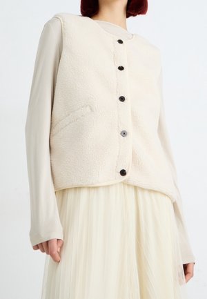 Persoon die een crèmekleurige fleece gilet met knopen draagt over een beige shirt met lange mouwen en een geplooide gebroken witte rok, staand tegen een egale achtergrond.