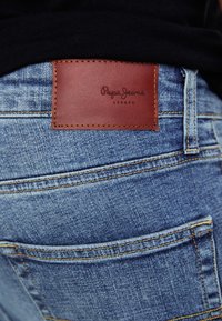 Plave traperice od denima sa smešnim kožnim oznakom s logotipom "Papa Jeans London." Vidljive šavove i džepovi doprinose dizajnerskim detaljima.